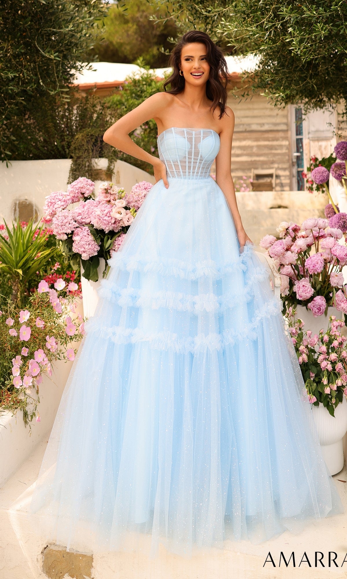 A woman wearing the Amarra Strapless Sheer-Bodice Prom Ball Gown 88794 with a glitter-tulle overlay stands outside near pink flowers and greenery, smiling at the camera.